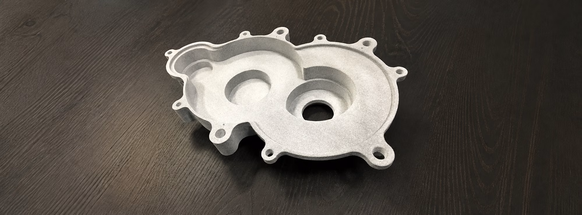 Light gray MJF housing with two circular cavities, recessed channels, and perimeter mounting tabs on a dark wood surface